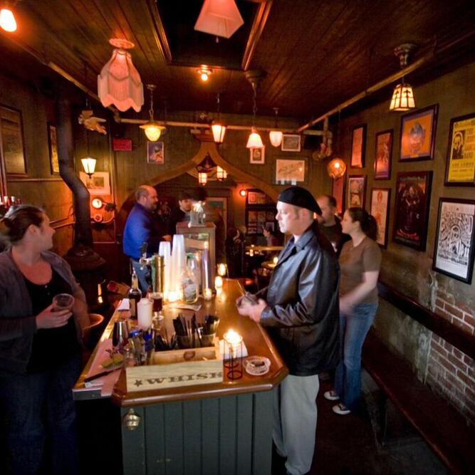 A small group of people gather in a small, dimly lit bar with a wood burning stove and unique decor