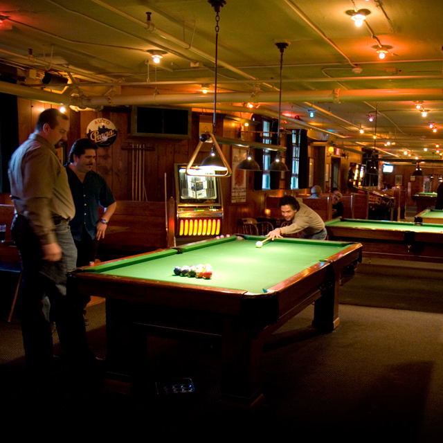 1 man shoots pool in dimly lit bar while 2 men watch