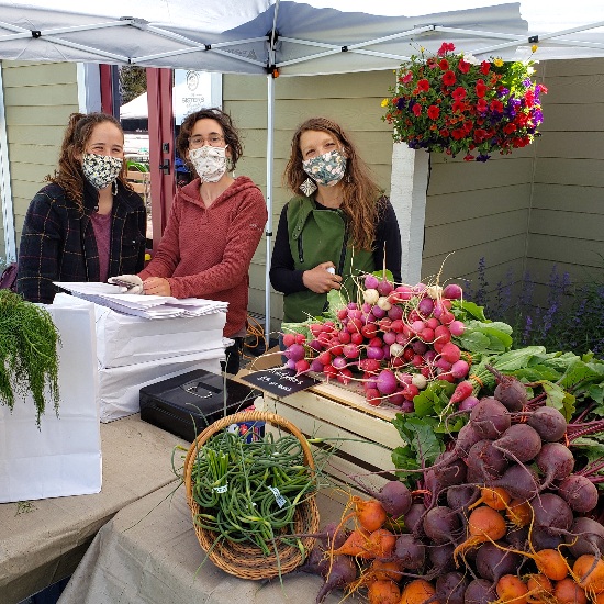 three people shown with masks on in front of produce stand serving radishes, beets and garlic scapes