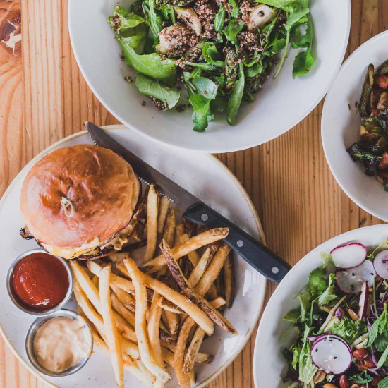 multiple plates of food shown on a wooden table including, a cheese burger and fries and an arugula salad with quinoa