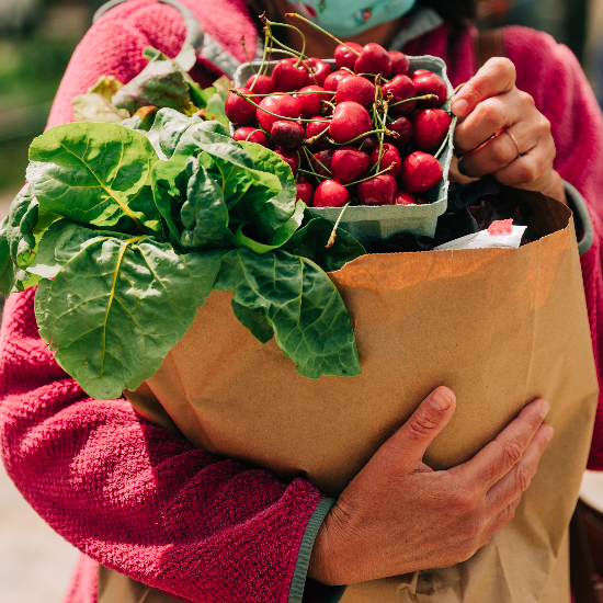 person shown holding brown bag full of produce with swiss chard and red cherries on top