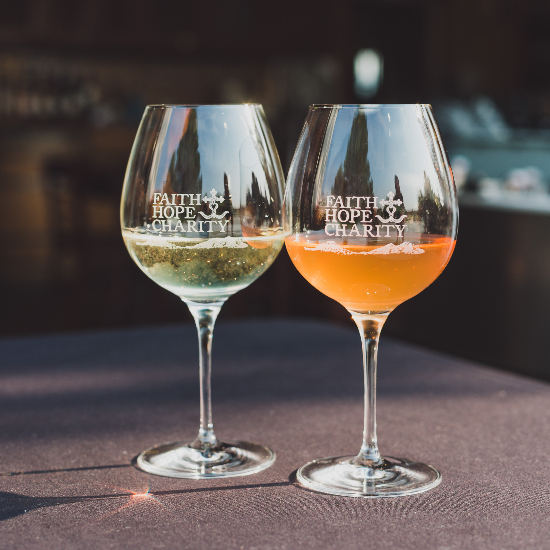 two glasses of wine, one white one orange with the faith hope and charity logo on the front of the glass