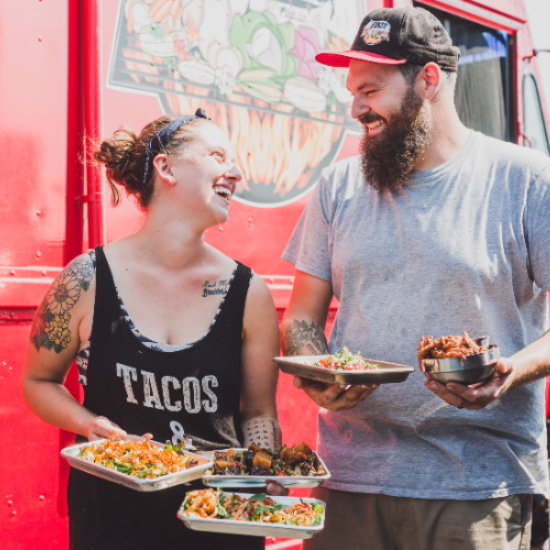 Owners of Feast Food Co. Chris and Emma standing in front of their food truck with plates of food laughing