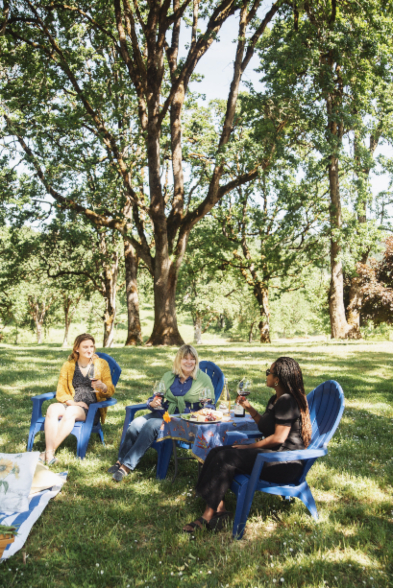 Three women enjoying food and wine in a grassy area under some trees.