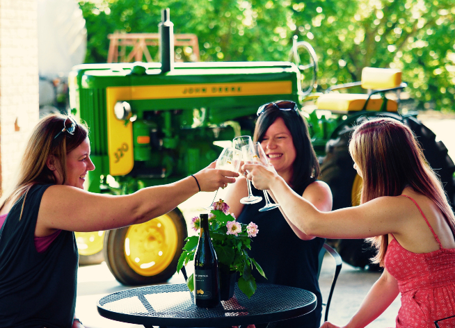 Three women drinking wine with a green tractor in the background.