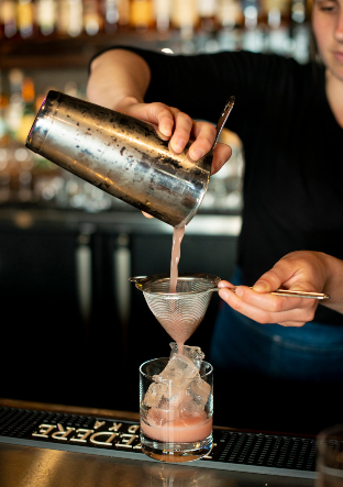 A bartender pouring a cocktail into a glass with ice.