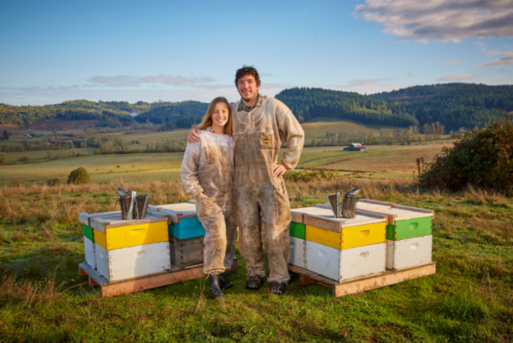 Two people standing in front of beehives with a scenic background.