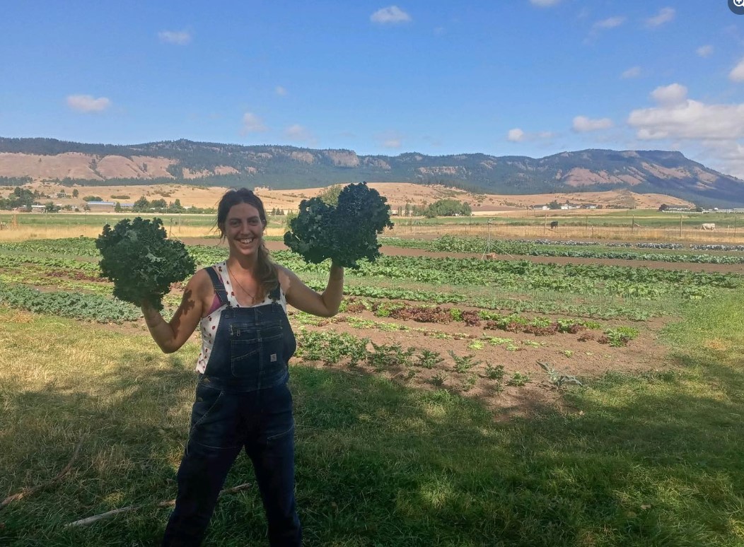 farmer poses holding a bunch of greens in each hand with scenic background of hills and blue sky