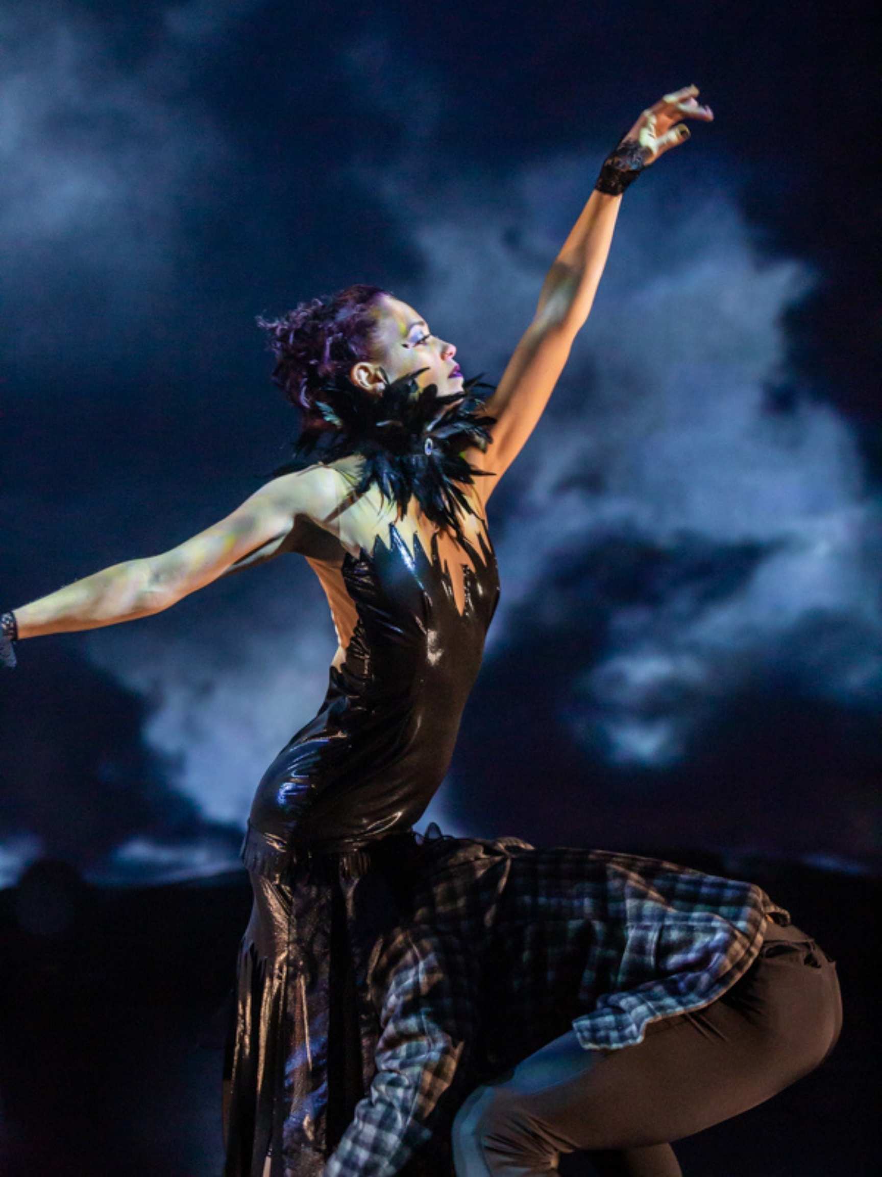 Dancer in black feather dress reaches for sky with night clouds in background