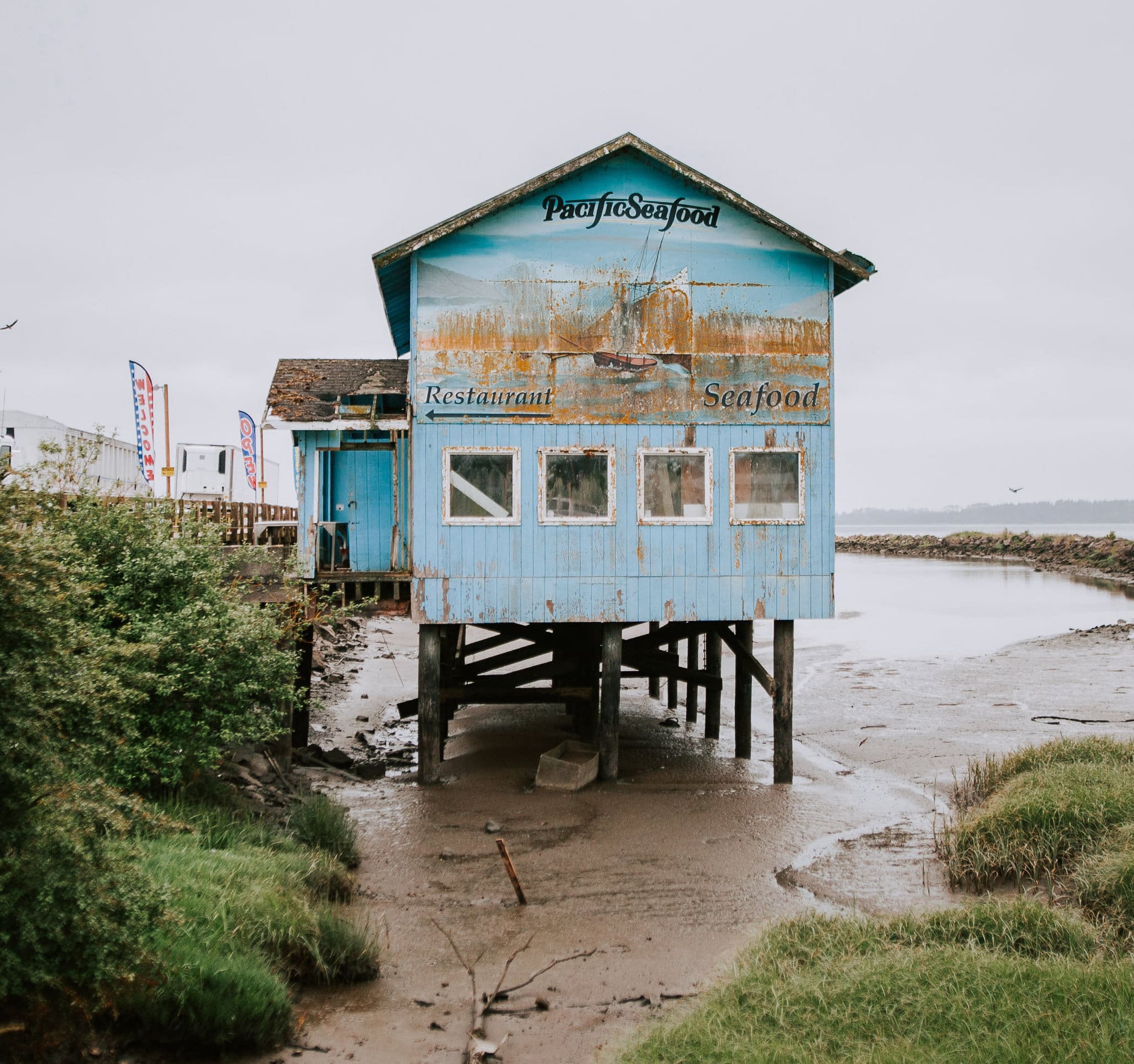 A blue building that reads Pacific Seafood sits on the water at low tide, exposings its stilts.
