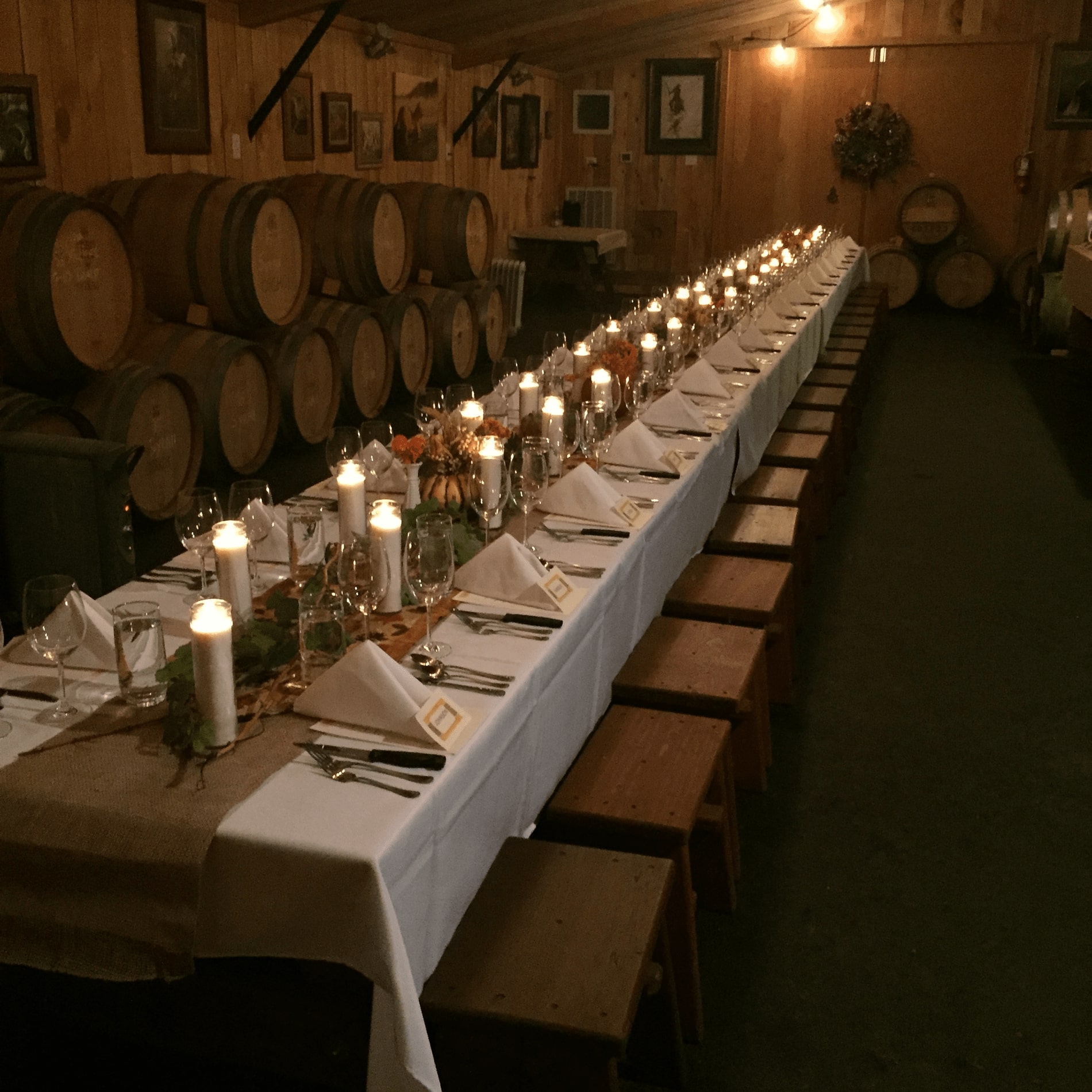 Image of long table settings with candles, napkins all in a row. A dimly lit room with barrels against the wall.