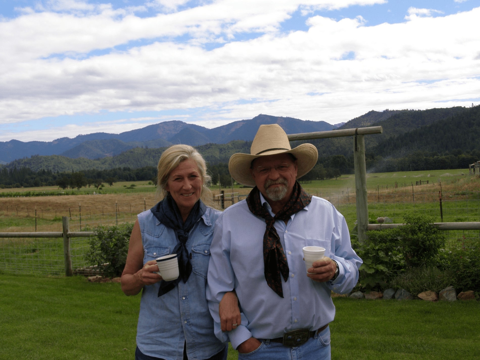 Plaisance Ranch owners standing infront of corral with mountain scape and clouds.