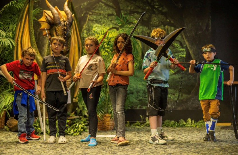 A group of kids lined up holding various props while standing in front of a backdrop with a dragon in the forest