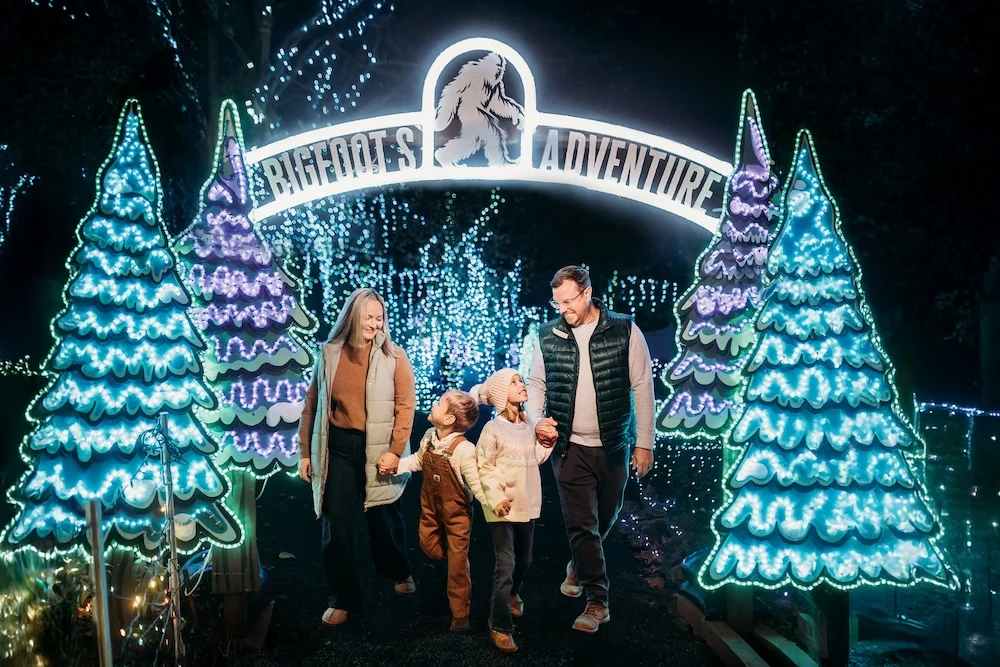 A family with two young kids hold hands while walking down a path decorated with christmas tree cutouts and colorful lights