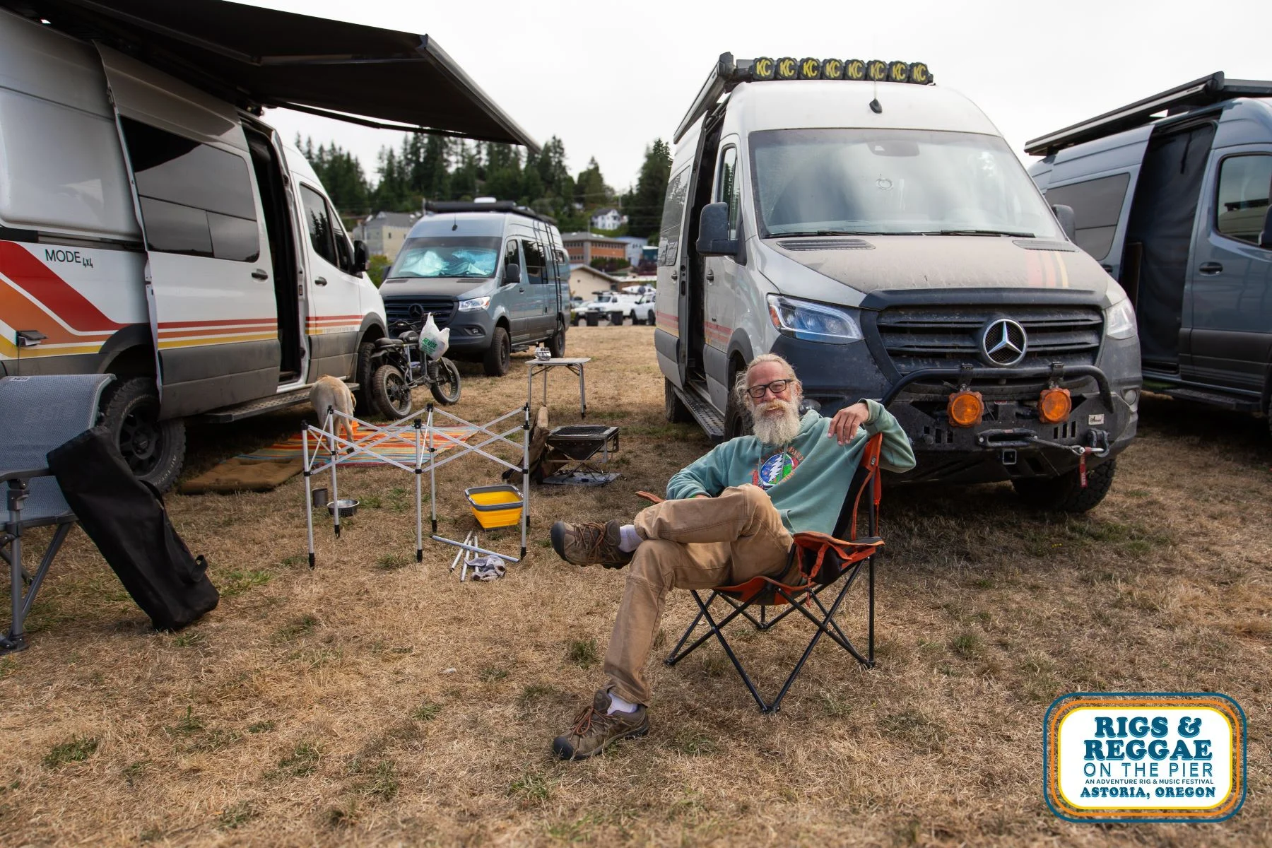 Man with a white beard lounges in camp chair with campervans around him in a field