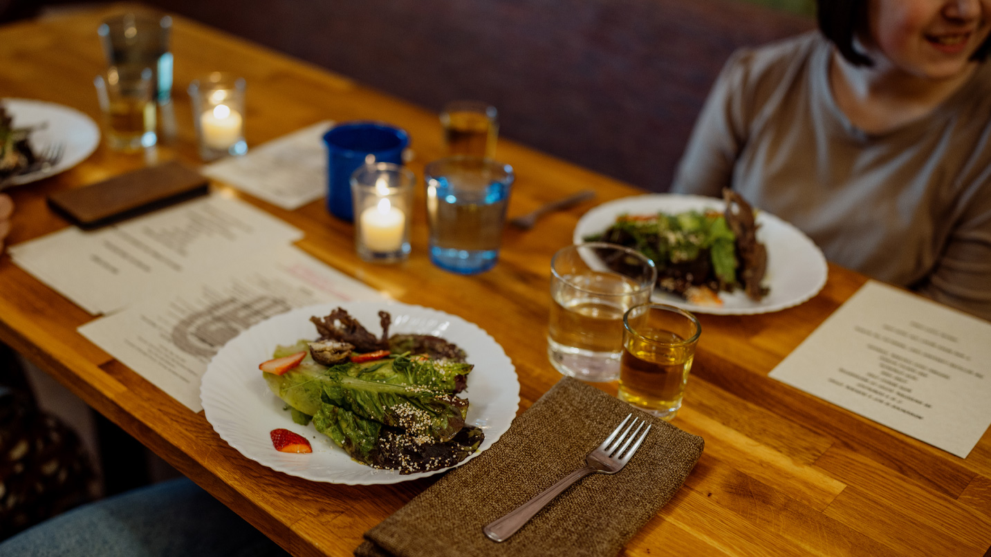 A wooden dining table with menus, drinking glasses, candles and white plates with salad greens