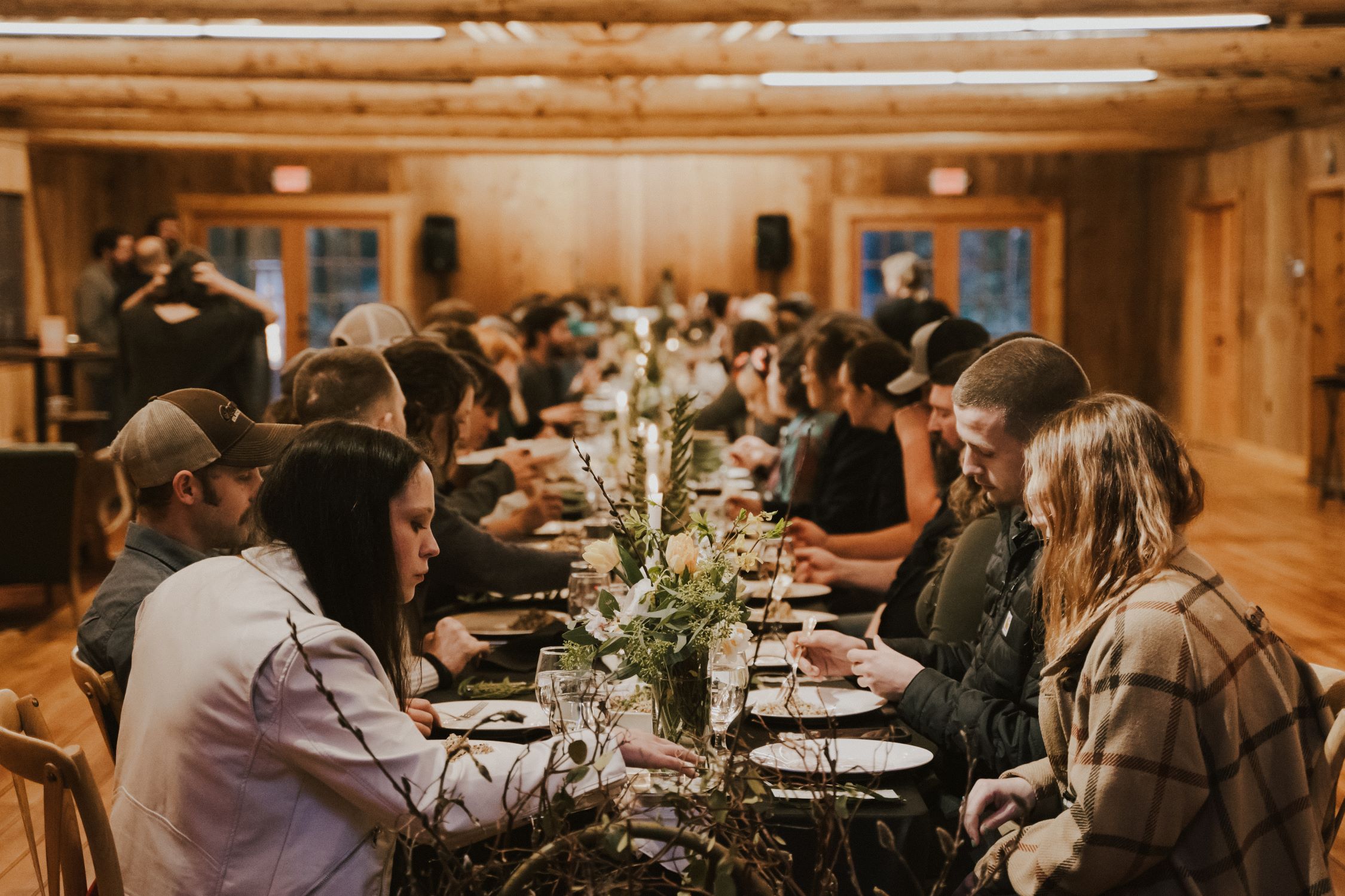 A group of diners sit at a long table adorned with greenery inside a log cabin style building