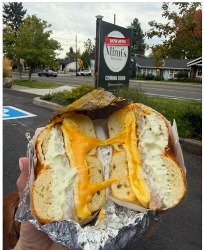 A person holds a bagel sandwich with egg and cheese outside in front of a business sign for Mimi's Bagel Deli