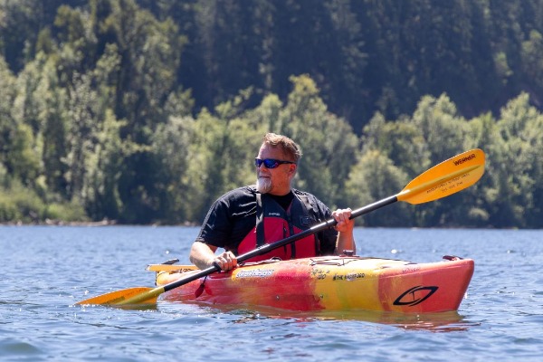 A man in sunglasses and a black tshirt paddles in an orange and yellow kayak on a lake surrounded by trees