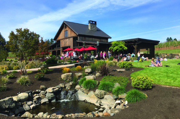 A barn-like winery with clean landscaping and groups of people sitting in the grass on a blue sky day