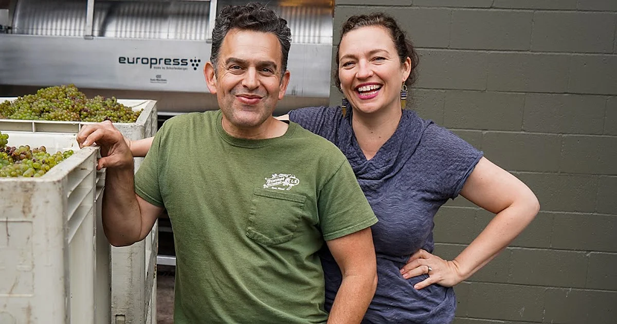 A man and a woman smile for at the camera standing next to large bins of wine grapes