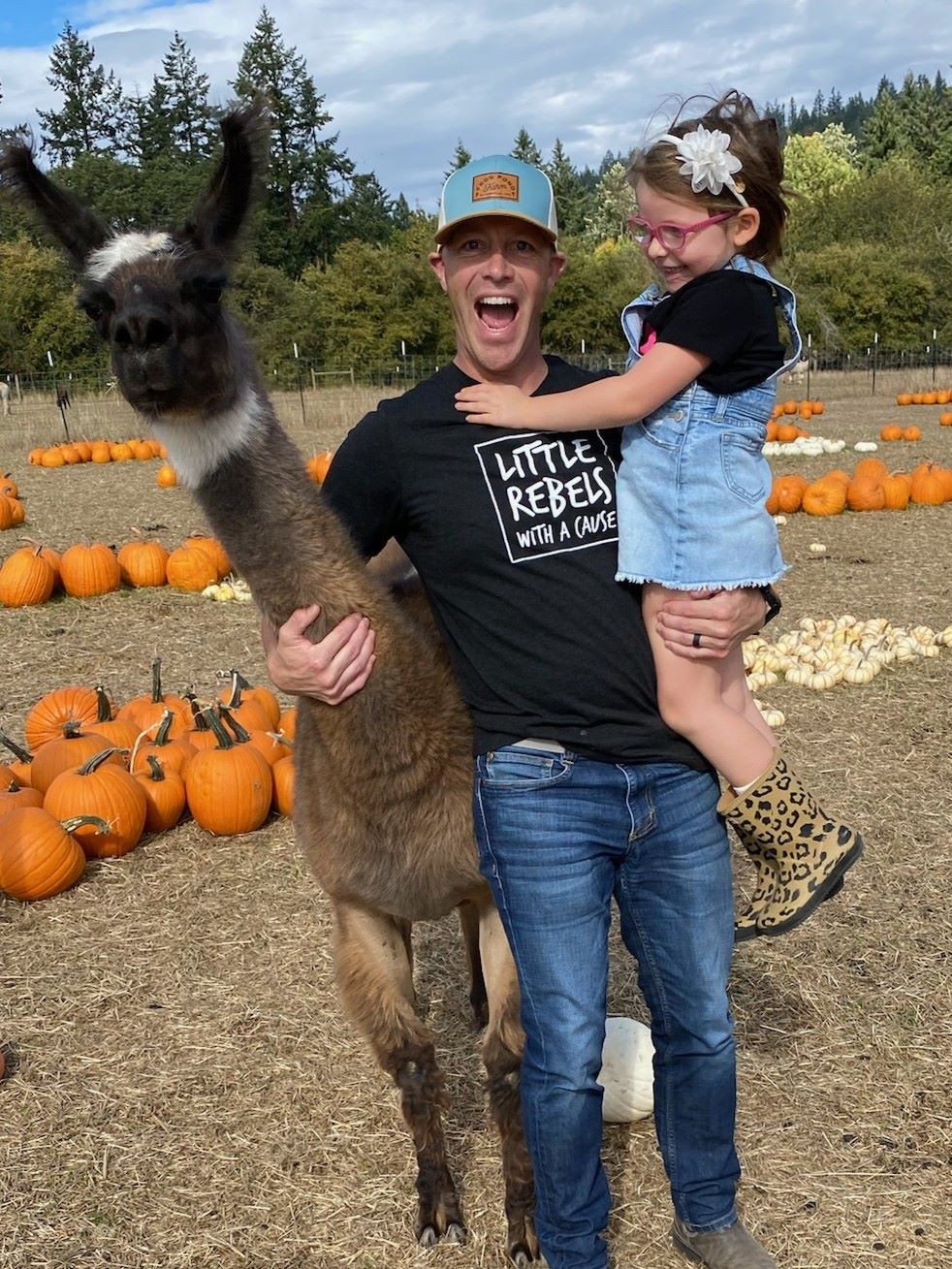 A smiling dad with their young daughter in one arm and a llama in the other smiles widely at the camera with piles of pumpkins in the background