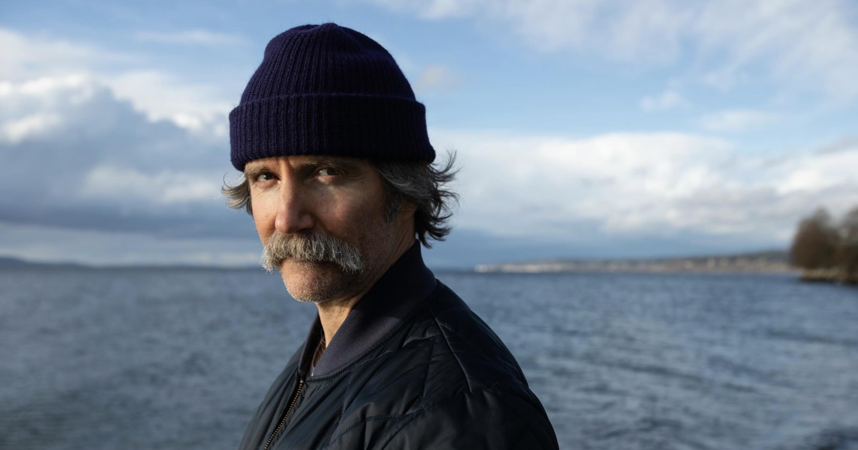 A man with a gray mustache wearing a dark beanie stands in front of a body of water