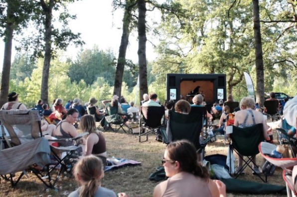 People sit in lawn chairs and on blankets outside surrounded by trees, facing a stage with musicians