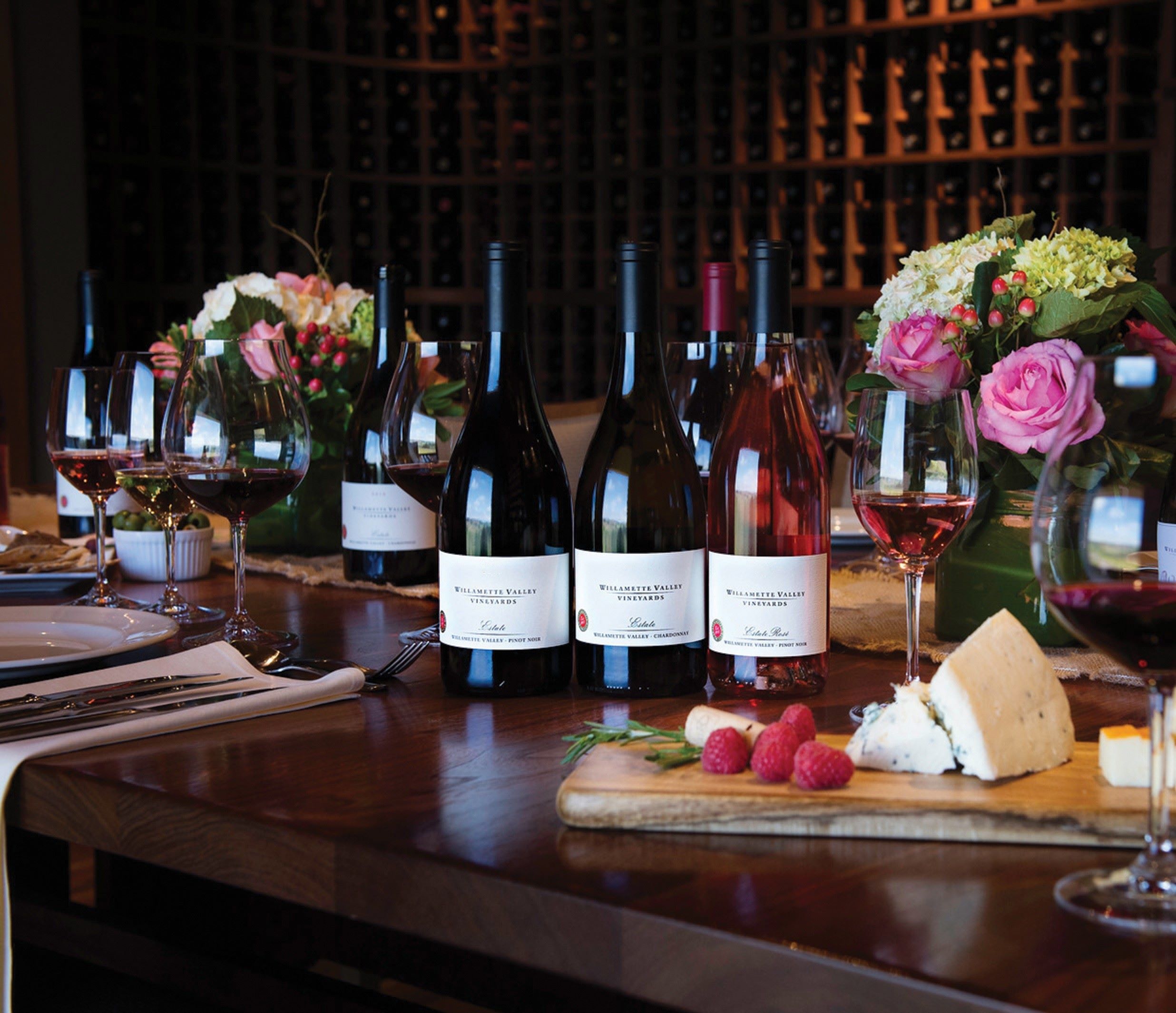Bottles of wine lined up on a bar with a cheese board and colorful bouquets of flowers