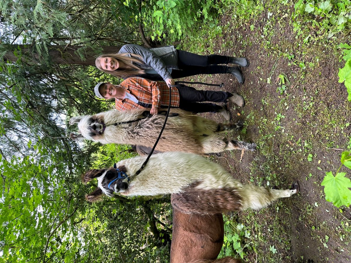 Two women and two llamas on a forested path smile at the camera