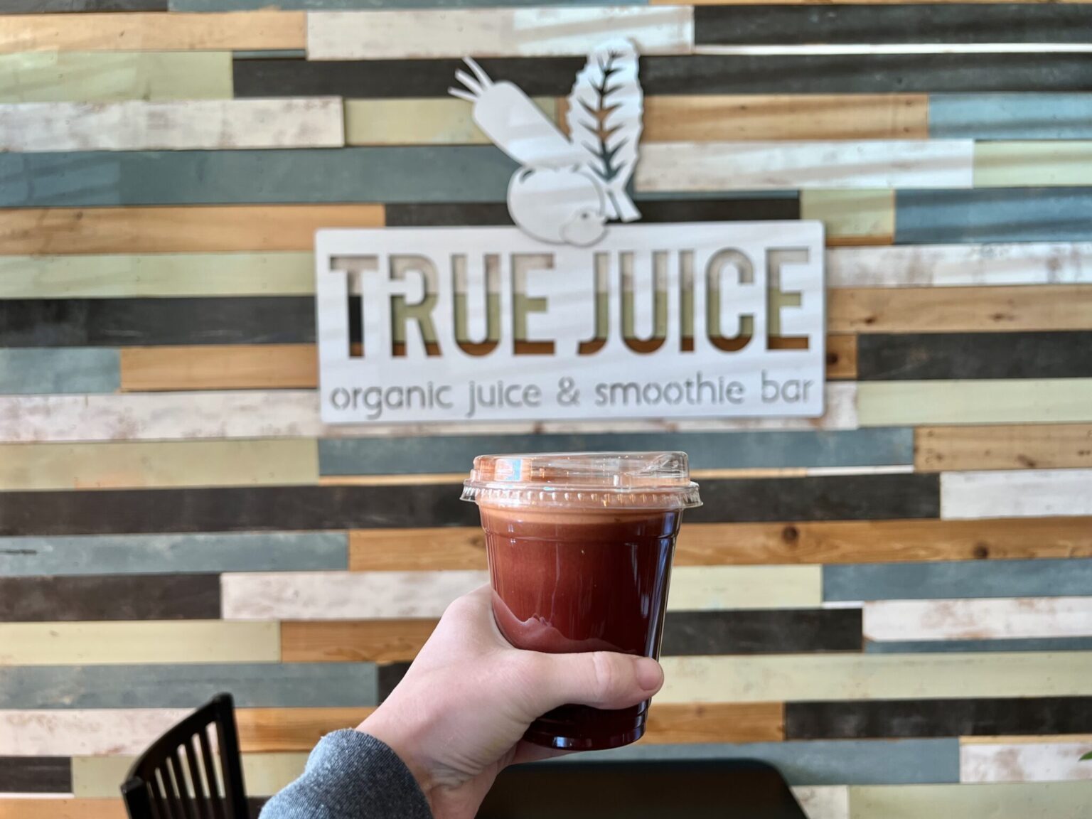 True Juice logo against a wall of colorful wooden planks with a smoothie being held in front