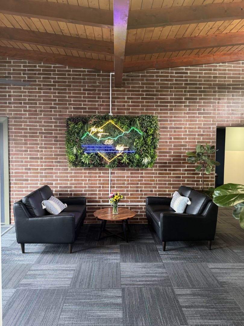 Cozy lounge space with black leather chairs, a round wooden coffee table, and a vibrant neon sign set into a vertical plant wall against exposed brick.