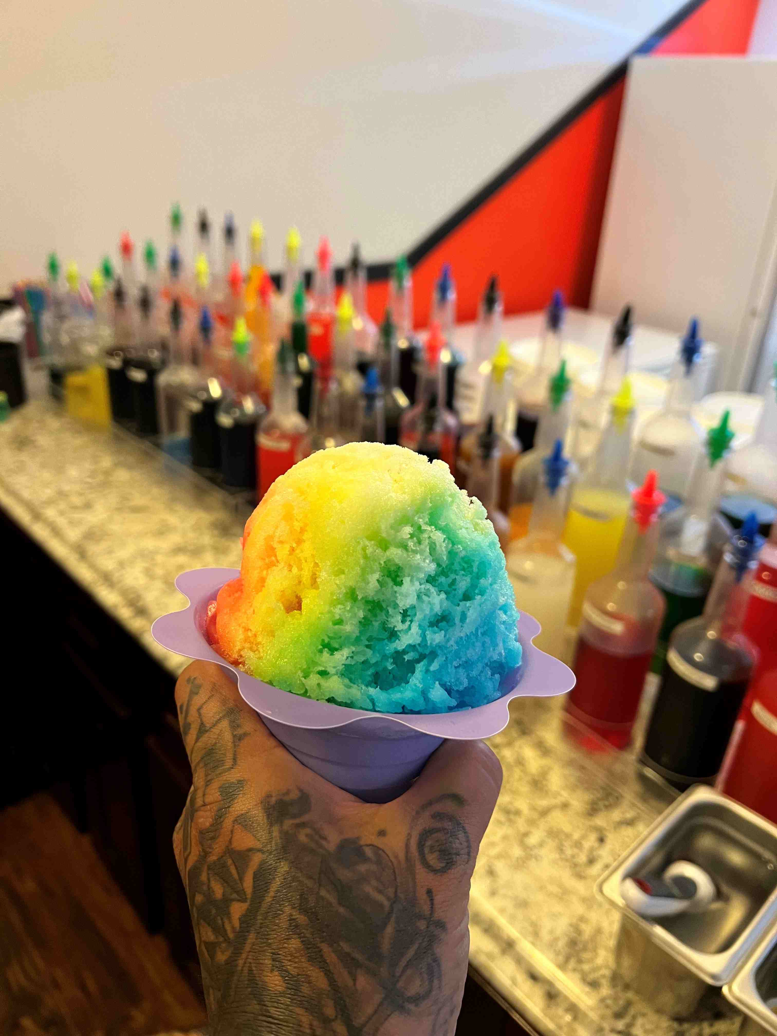 A tattoo covered hand holds up a rainbow colored shaved ice in front of bottles of favored syrup.