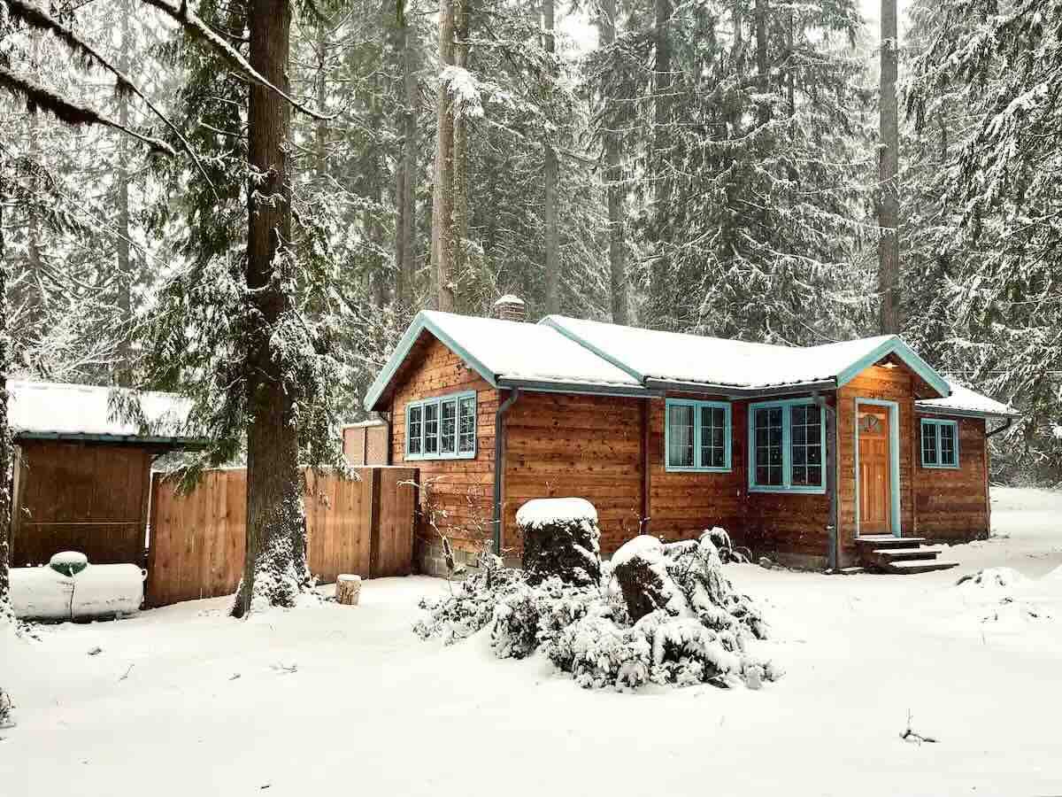 A cedar cabin with green trim sits in a snowy forest. Three steps lead up to the front door where the porch light is on.