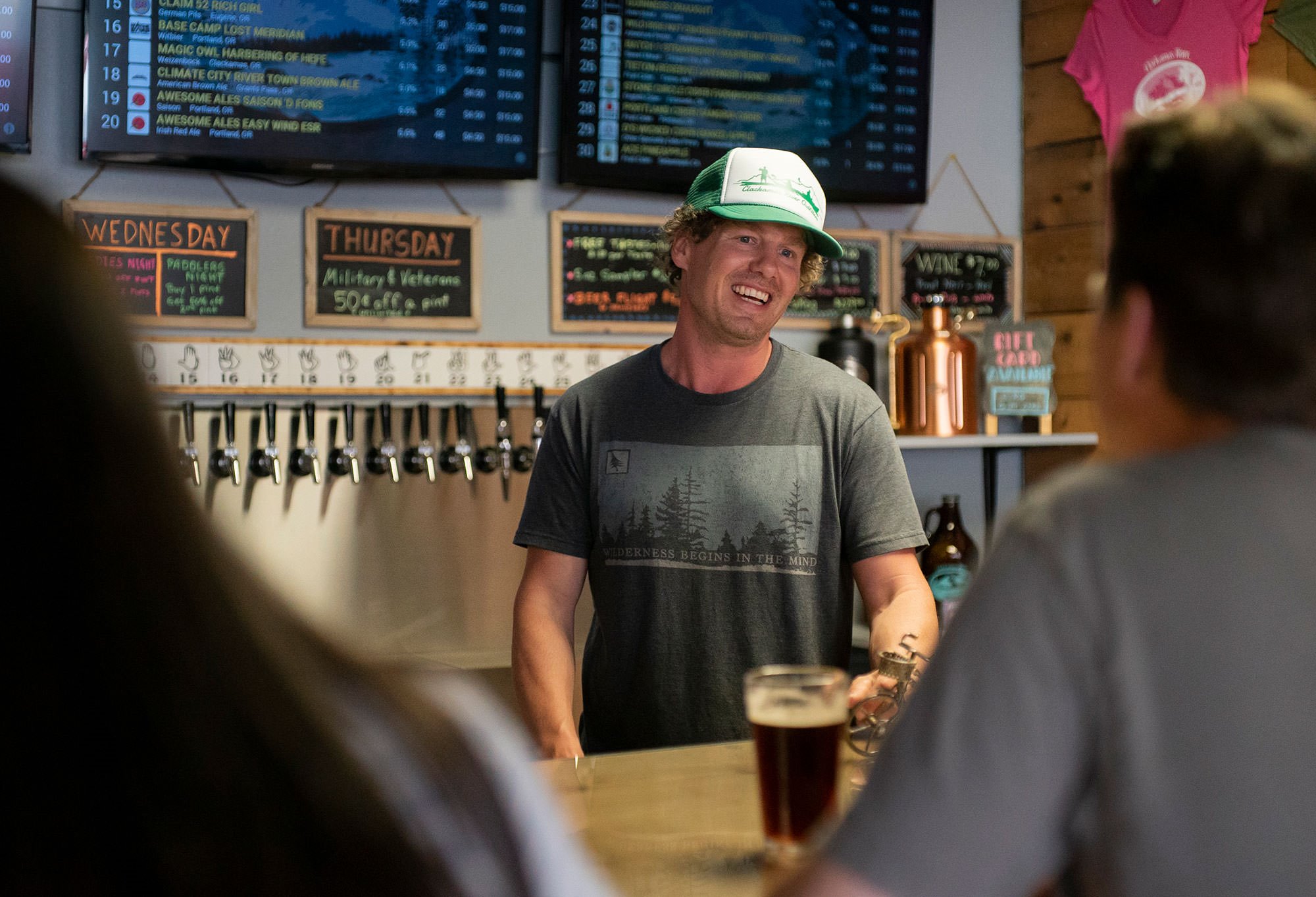 A bartender smiles while serving drinks at a lively bar with multiple beer taps and a menu displayed on screens above.