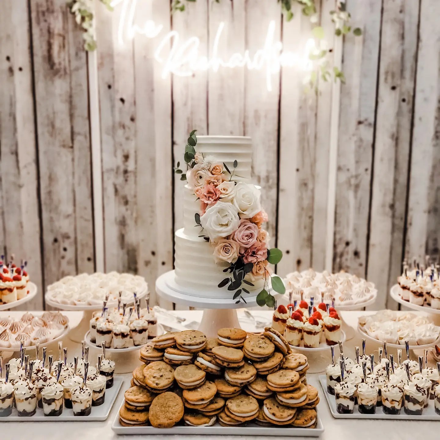 A white three tiered wedding cake with flowers sits in the center of a dessert table