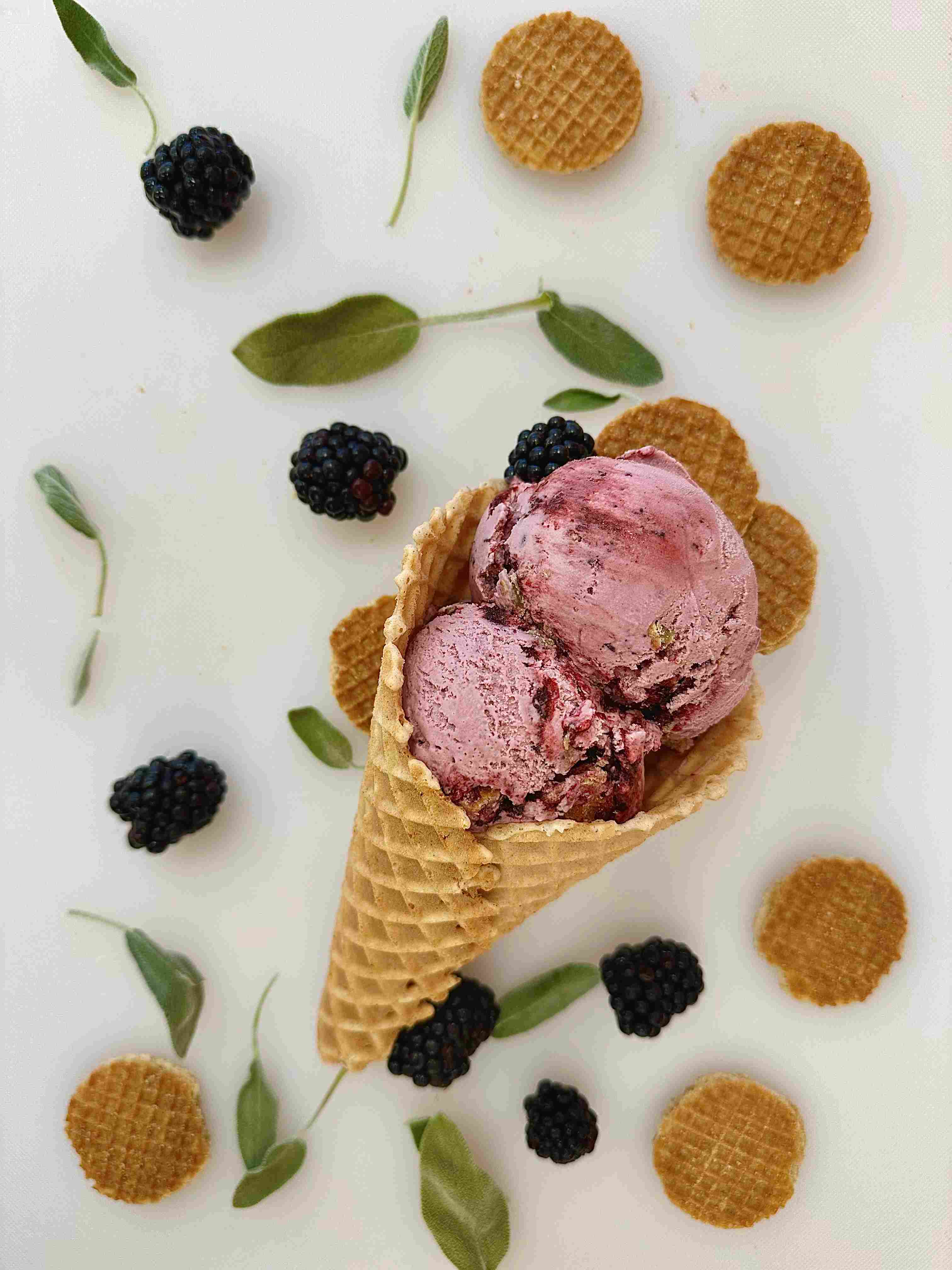 A two scoop ice cream cone lays on a counter surrounded by blackberries, wafer cookies and sage leaves.