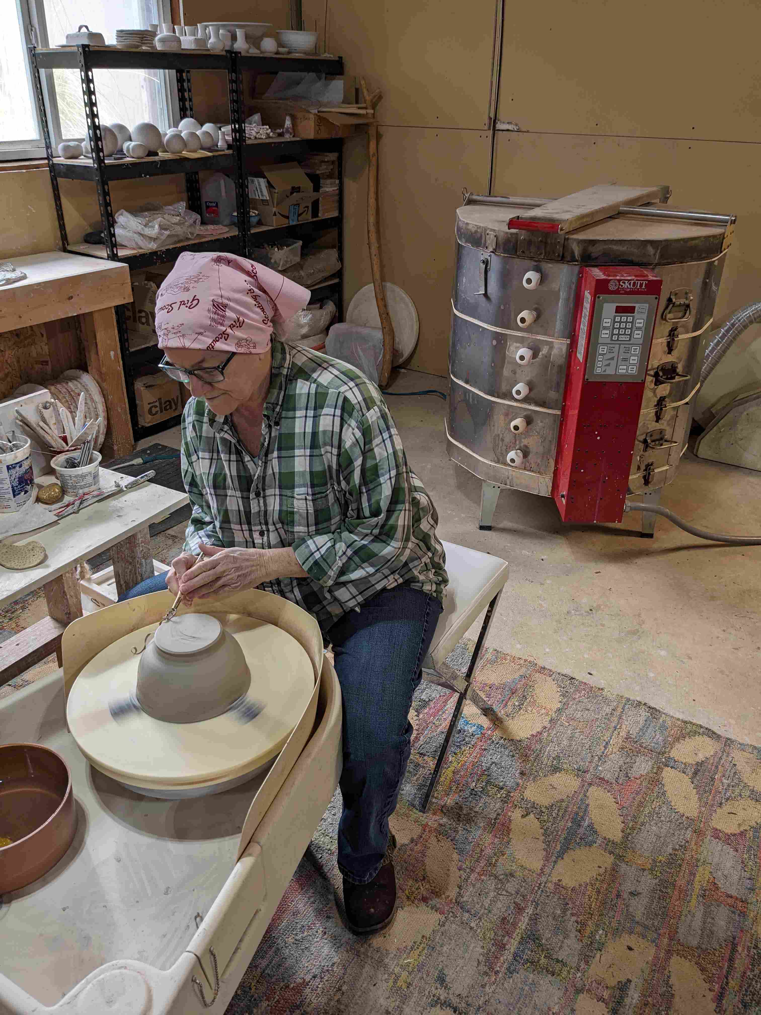 a ceramic artist wearing a pink bandana trims a clay bowl on a pottery wheel. Behind them is a ceramic kiln.