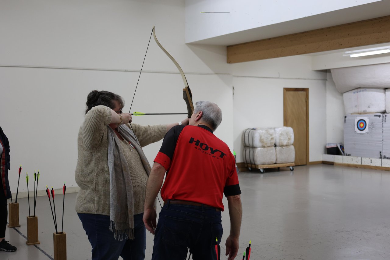 A student draws a bow while an instructor assists in an indoor archery range, with targets and arrows visible in the background.