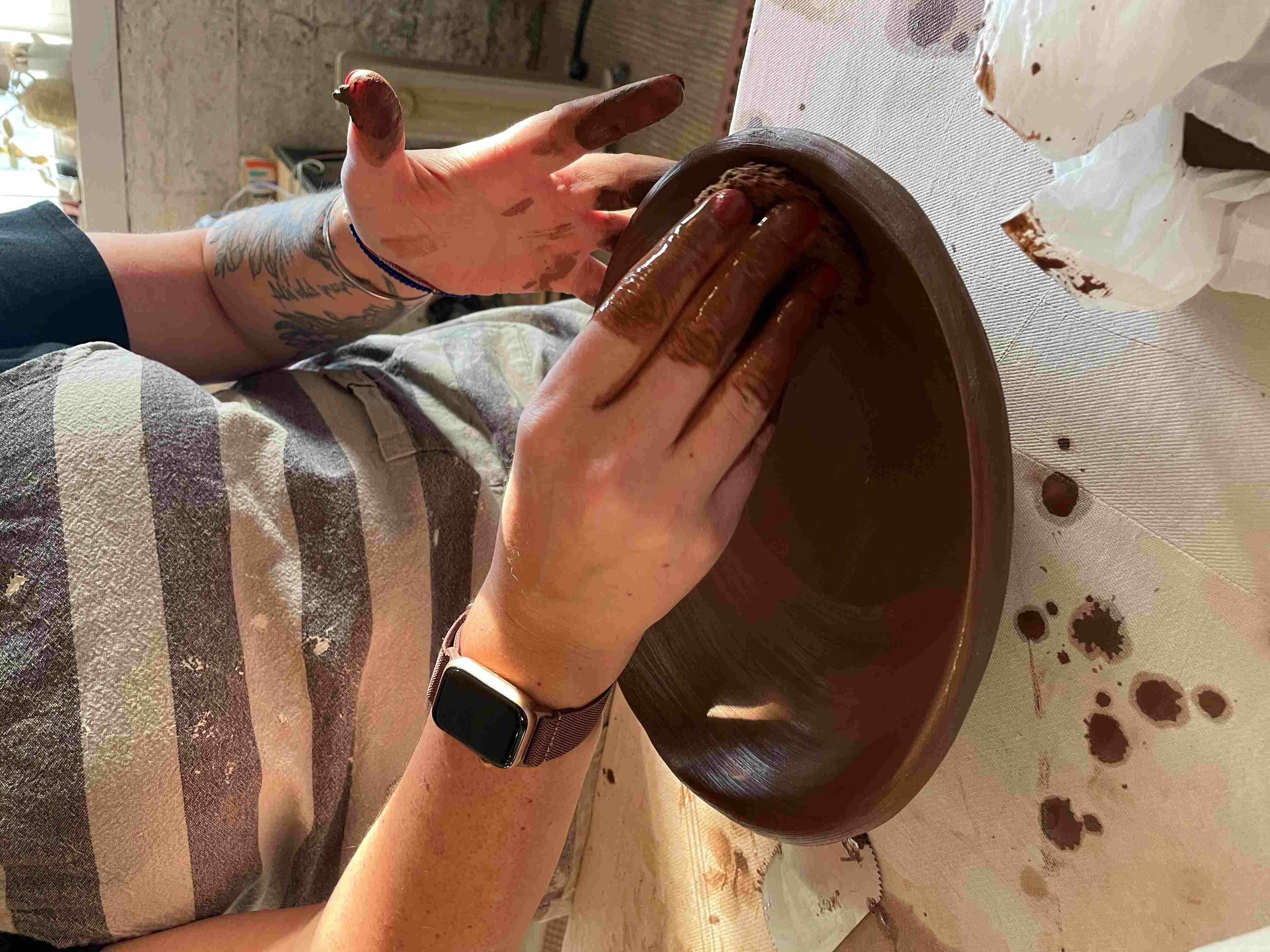 A potter in a grey & white striped shirt forms a ceramic bowl out of dark brown clay.