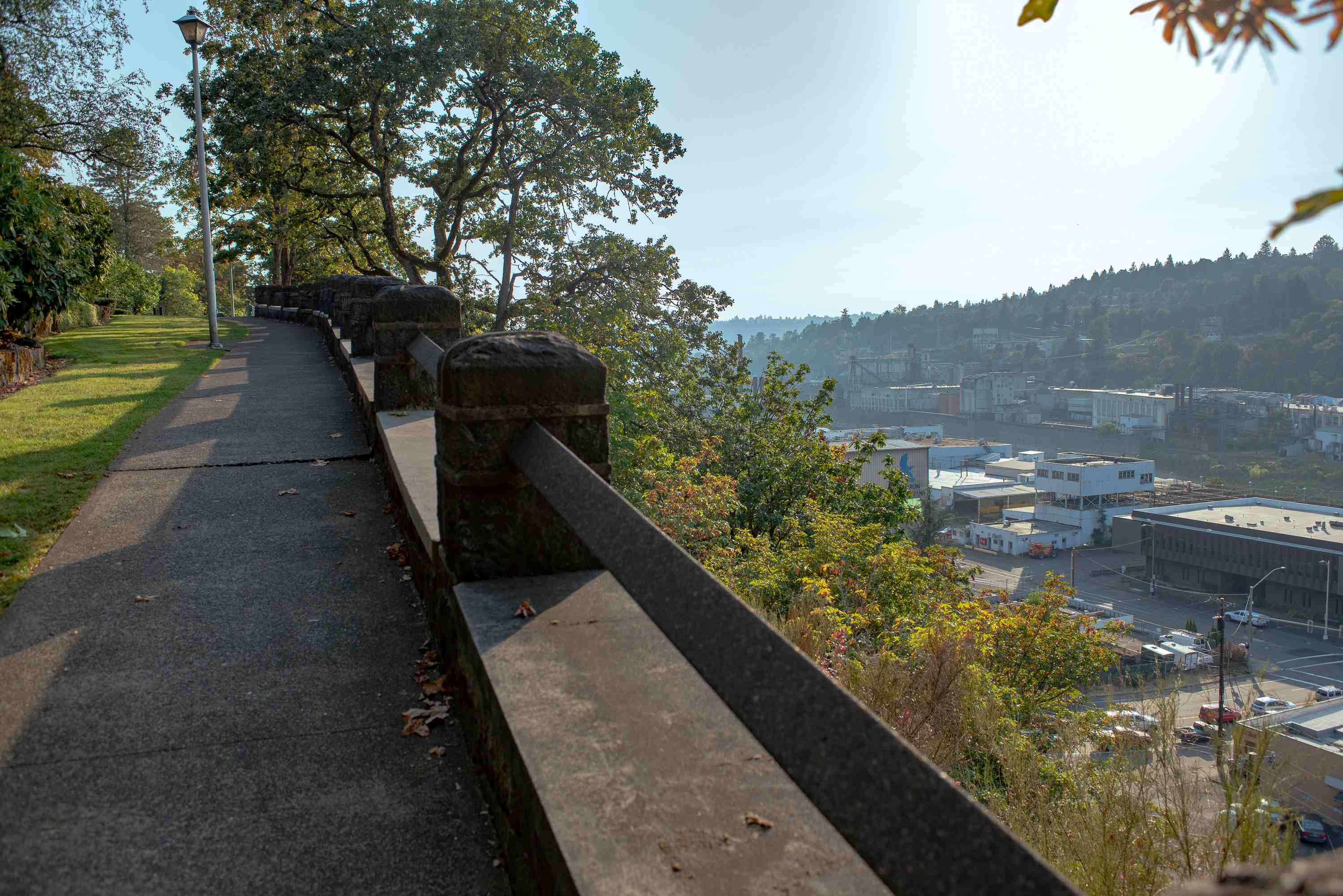 a paved path with grass to the left and bushes on the righthand slope overlook downtown Oregon City below