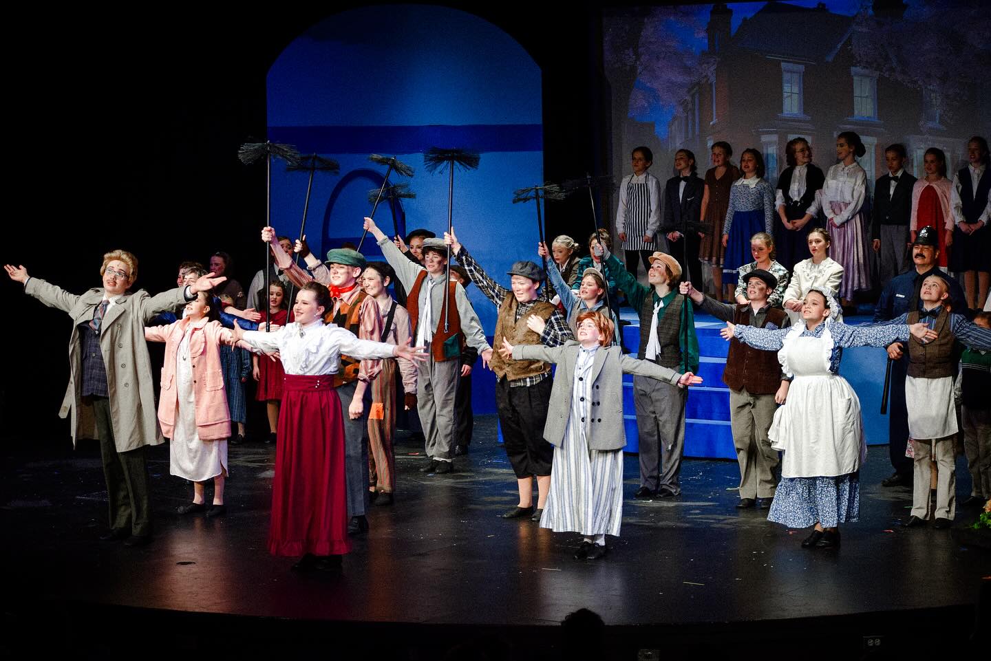 A lively stage performance featuring a large cast in period costumes, with actors holding props and striking dynamic poses. The backdrop shows a house, enhancing the theatrical setting.