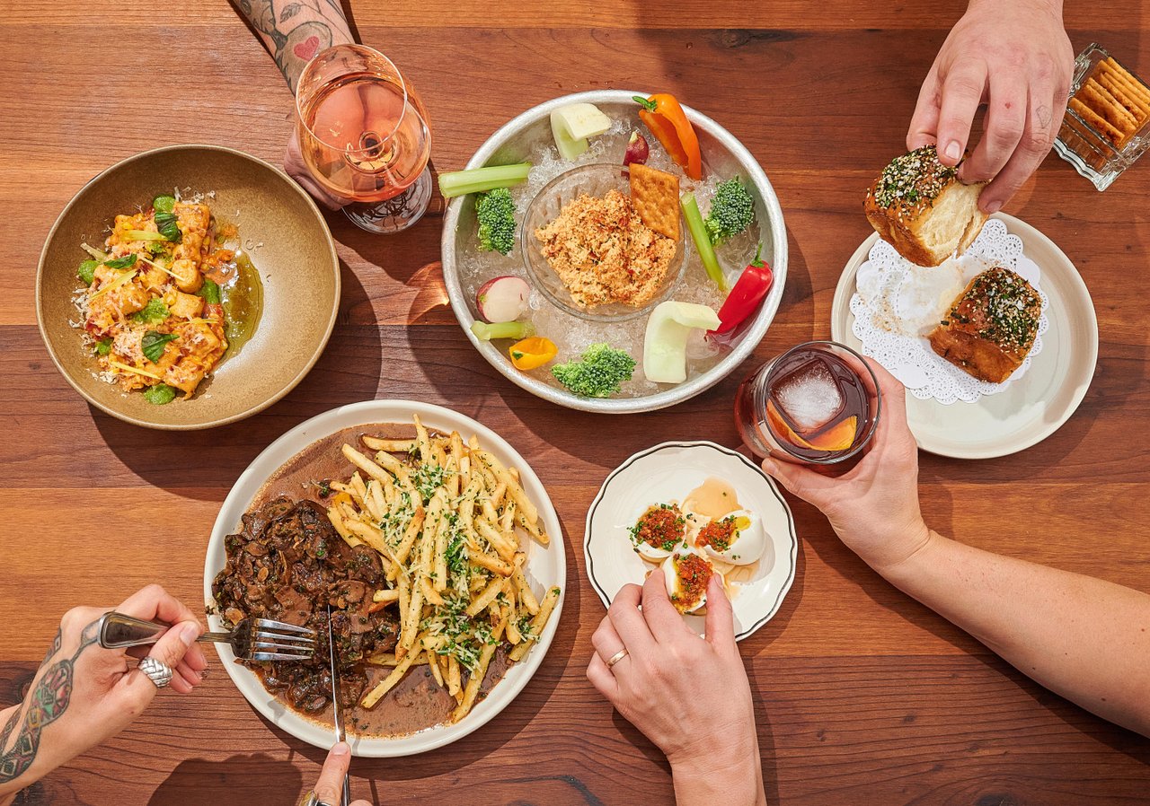 A wooden table displays a variety of dishes: pasta, seafood, and appetizers, alongside drinks. Hands reach for food and drinks, creating a lively dining scene.