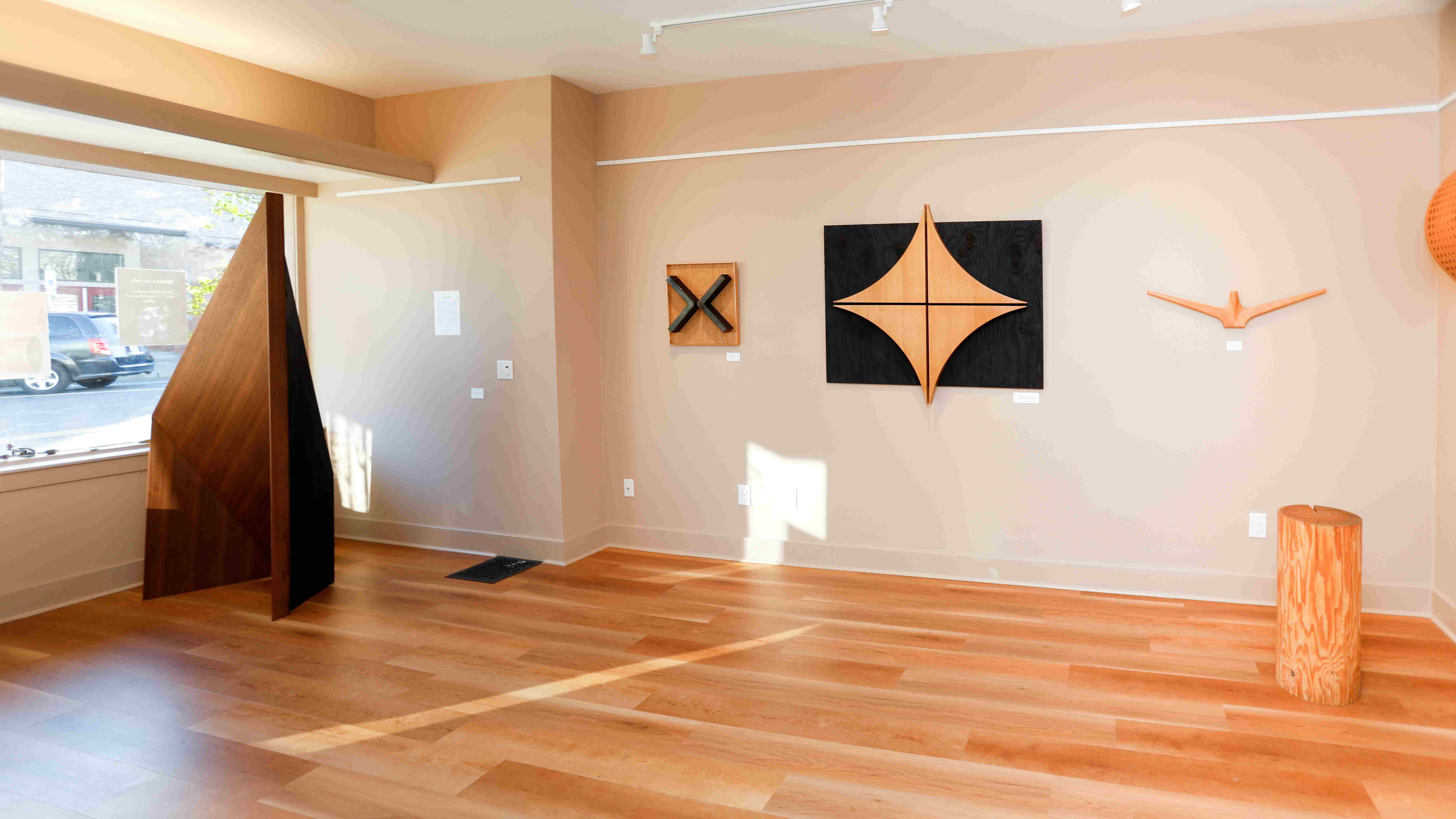 Interior of an art gallery featuring wooden sculptures and geometric wall art. Natural light illuminates the space, enhancing the warm wood tones.