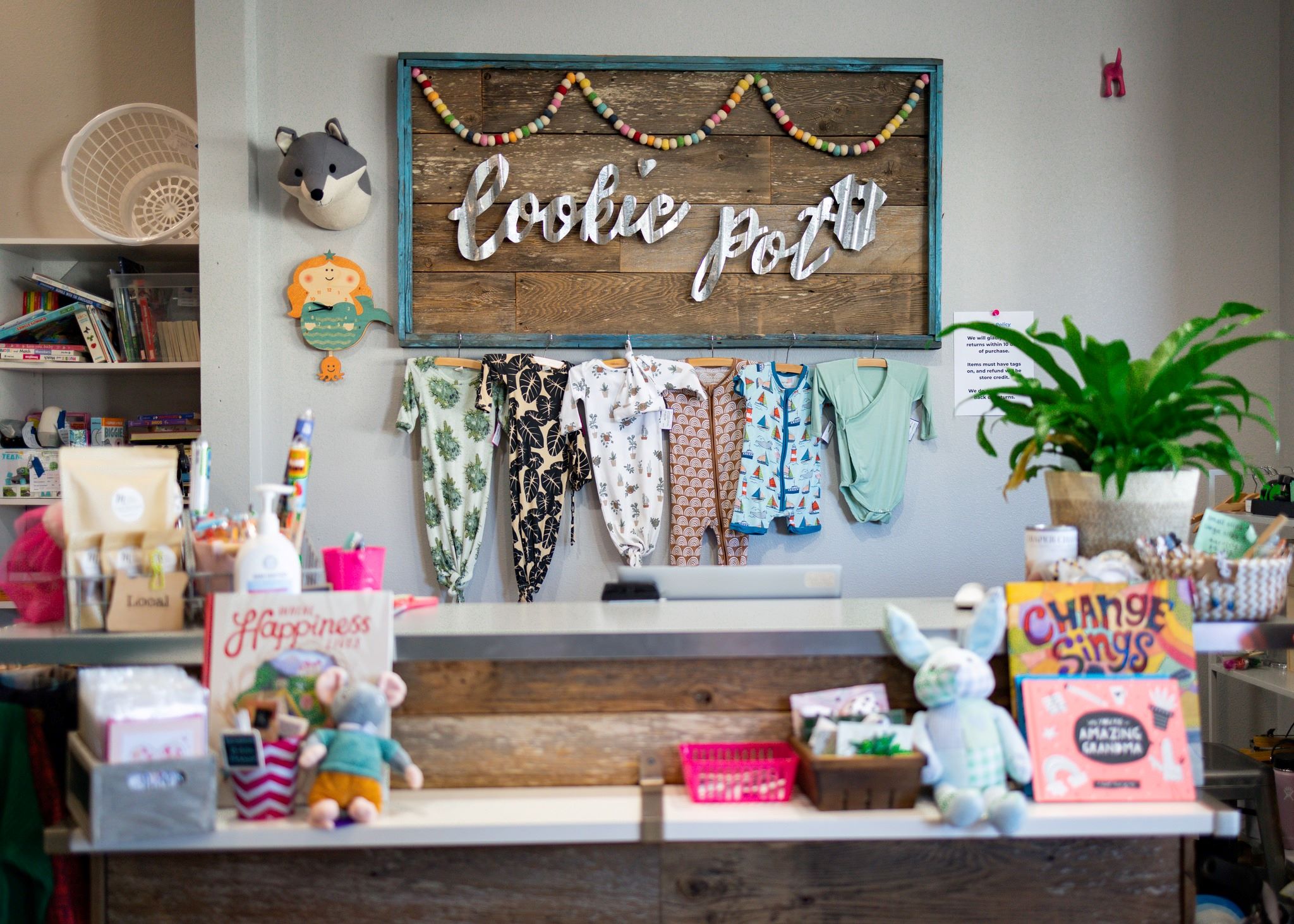 Infant clothing hangs behind a counter & point of sale. Wooden & Metal sign reads Cookie Pot