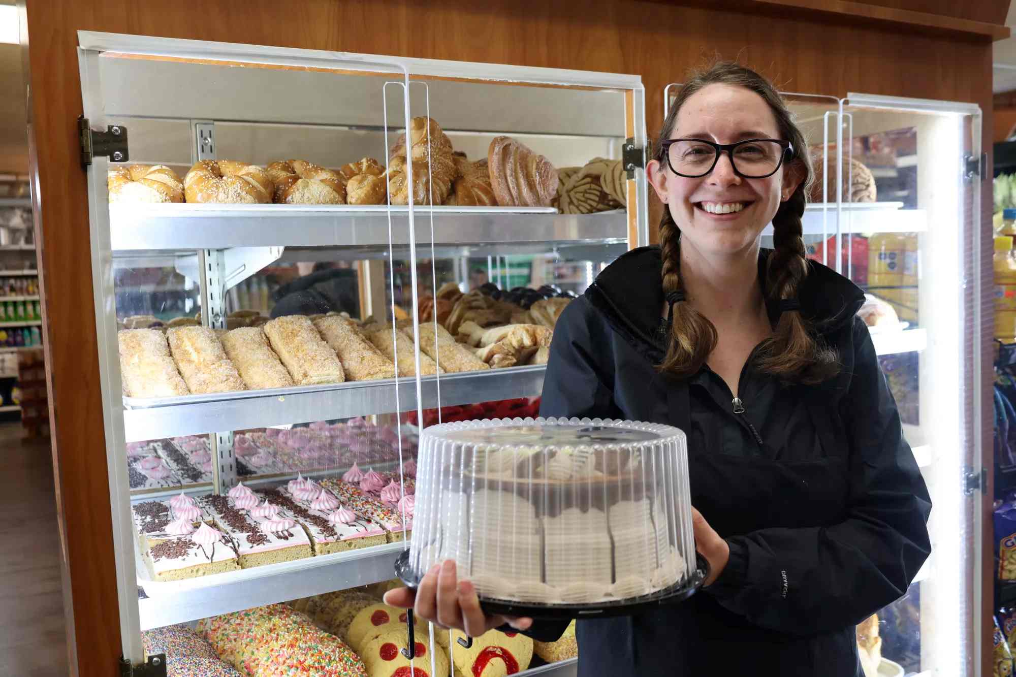 Shopkeeper at La Conasuper smiles for the camera while holding a cake in front of the bakery case.