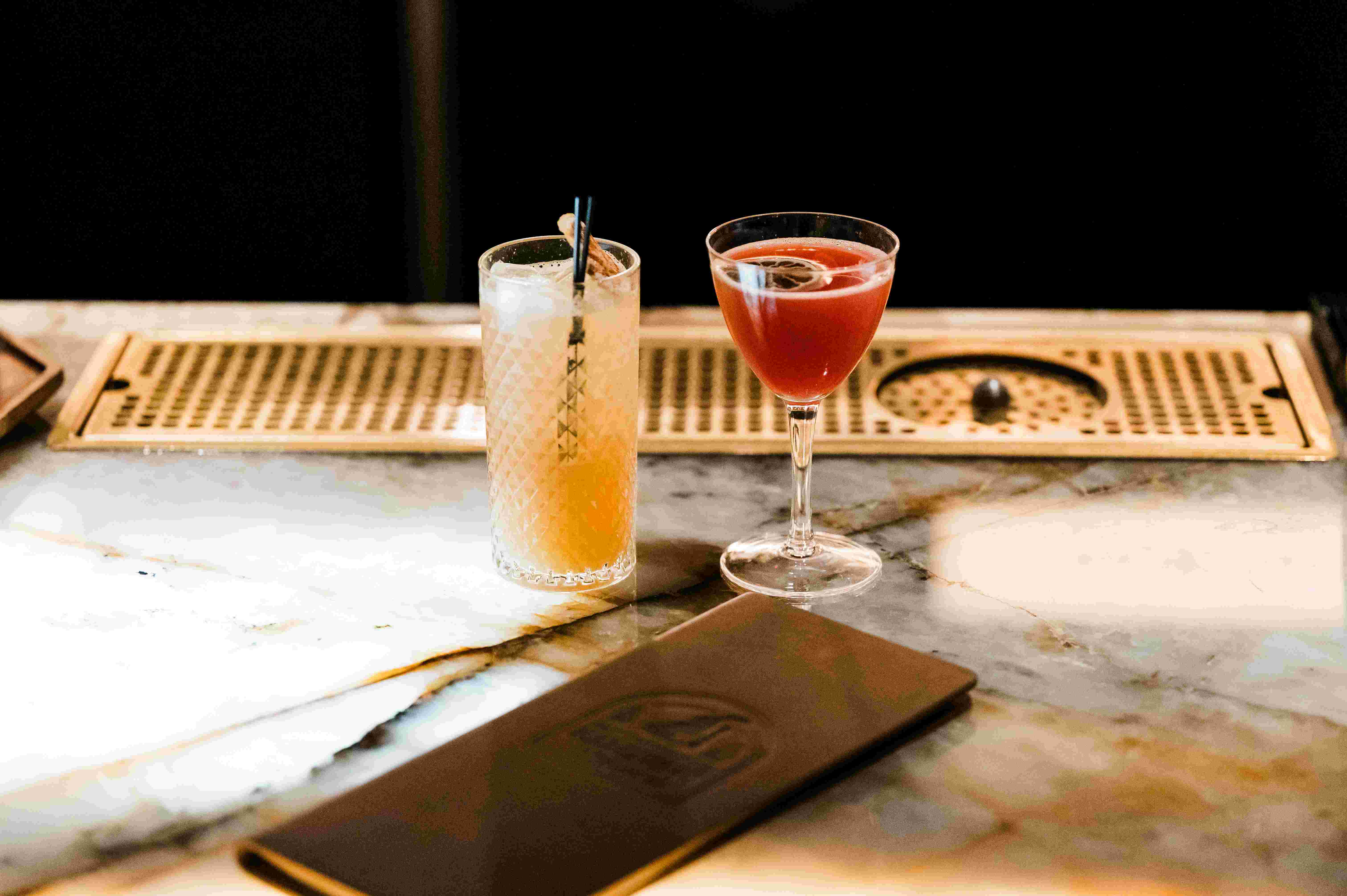 Two cocktails sit on a marble bar top: a tall glass with a straw and a short glass with a stem. A menu lies nearby.