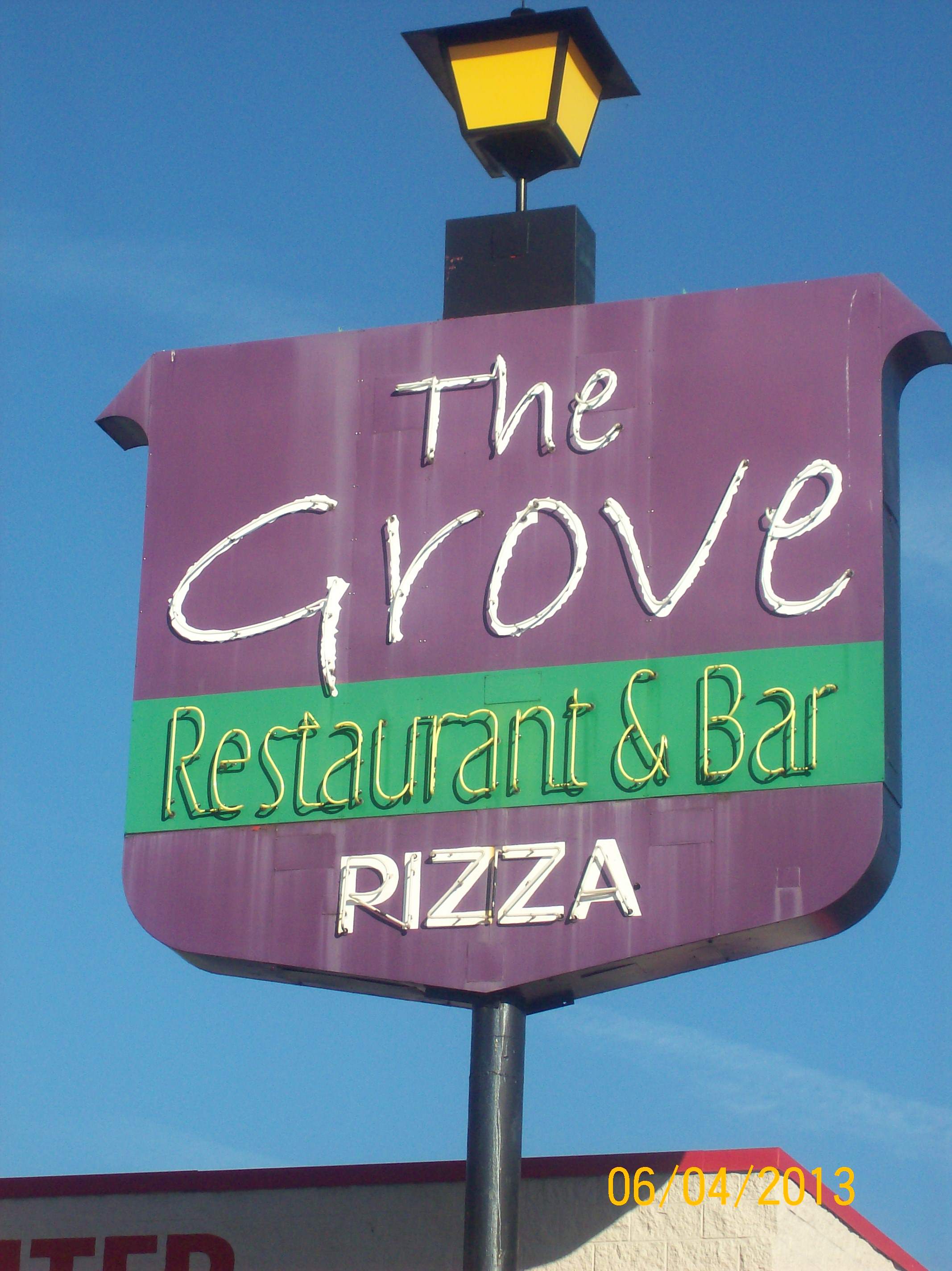 Street sign for The Grove Restaurant, Bar & Pizza stands against a blue sky. A lamp sits atop.