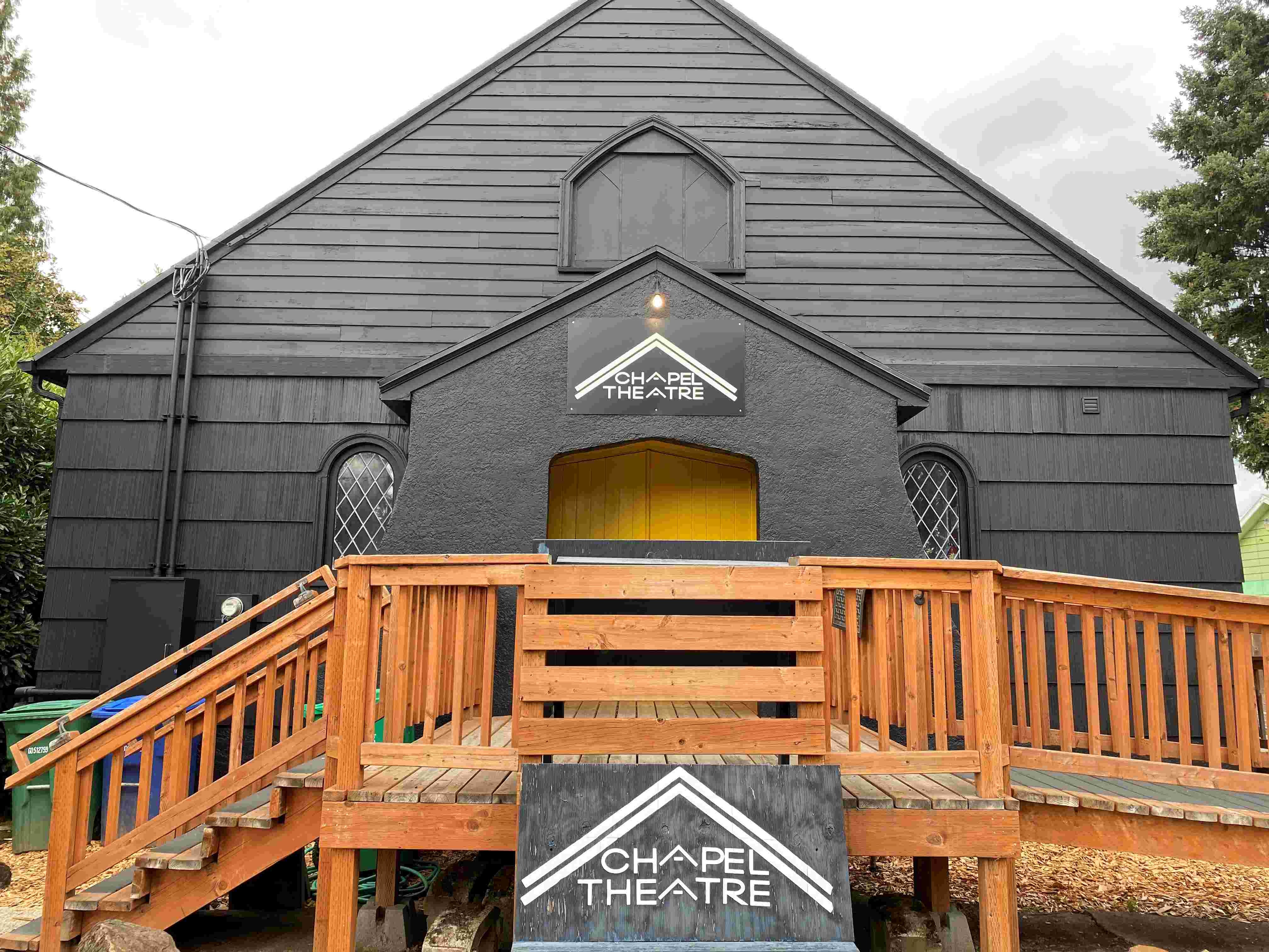 Black building with yellow door and cedar porch with ramp and stairs. Sign reads Chapel Theatre.