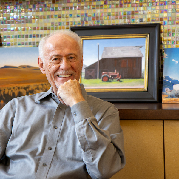 Photo of Gary Ernest Smith, a white man in his early eighties, seated in front of a painting of a red barn and tractor.
